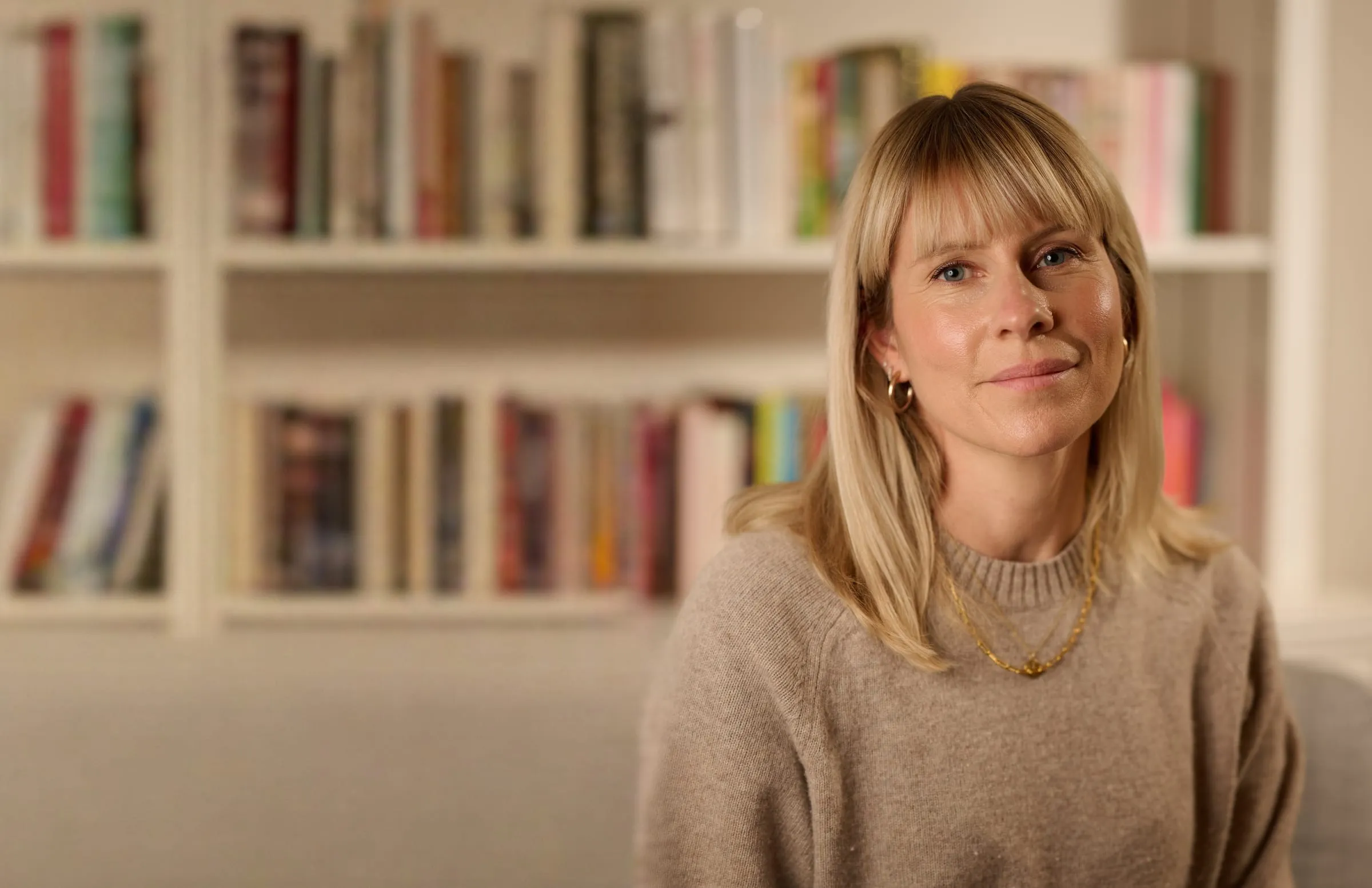 Alex Bickford, smiling, seated in front of a full bookshelf and wearing a soft beige jumper