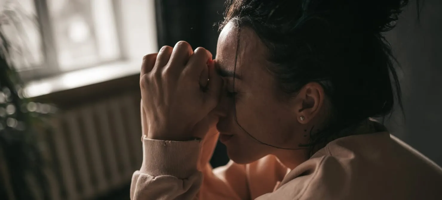 A woman sitting in soft window light, eyes closed, her hands pressed to her forehead
