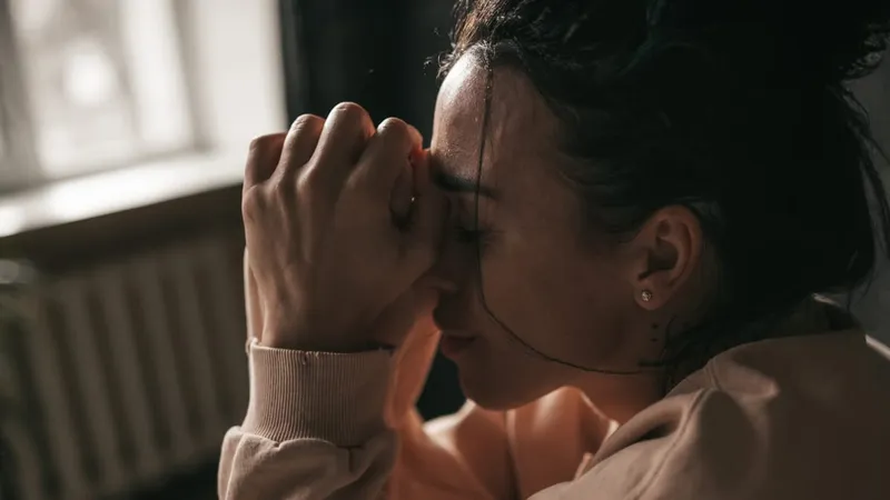 A woman sitting in soft window light, eyes closed, her hands pressed to her forehead