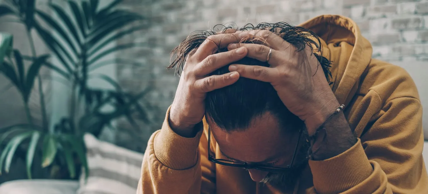 A man in a mustard hoodie sitting on a sofa, head bowed and both hands pressed to his head