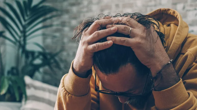 A man in a mustard hoodie sitting on a sofa, head bowed and both hands pressed to his head